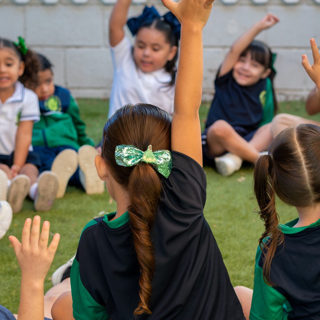 Alumnas y alumnos de preescolar de Colegio Encinos sentados en círculo sobre el pasto, levantando la mano para participar