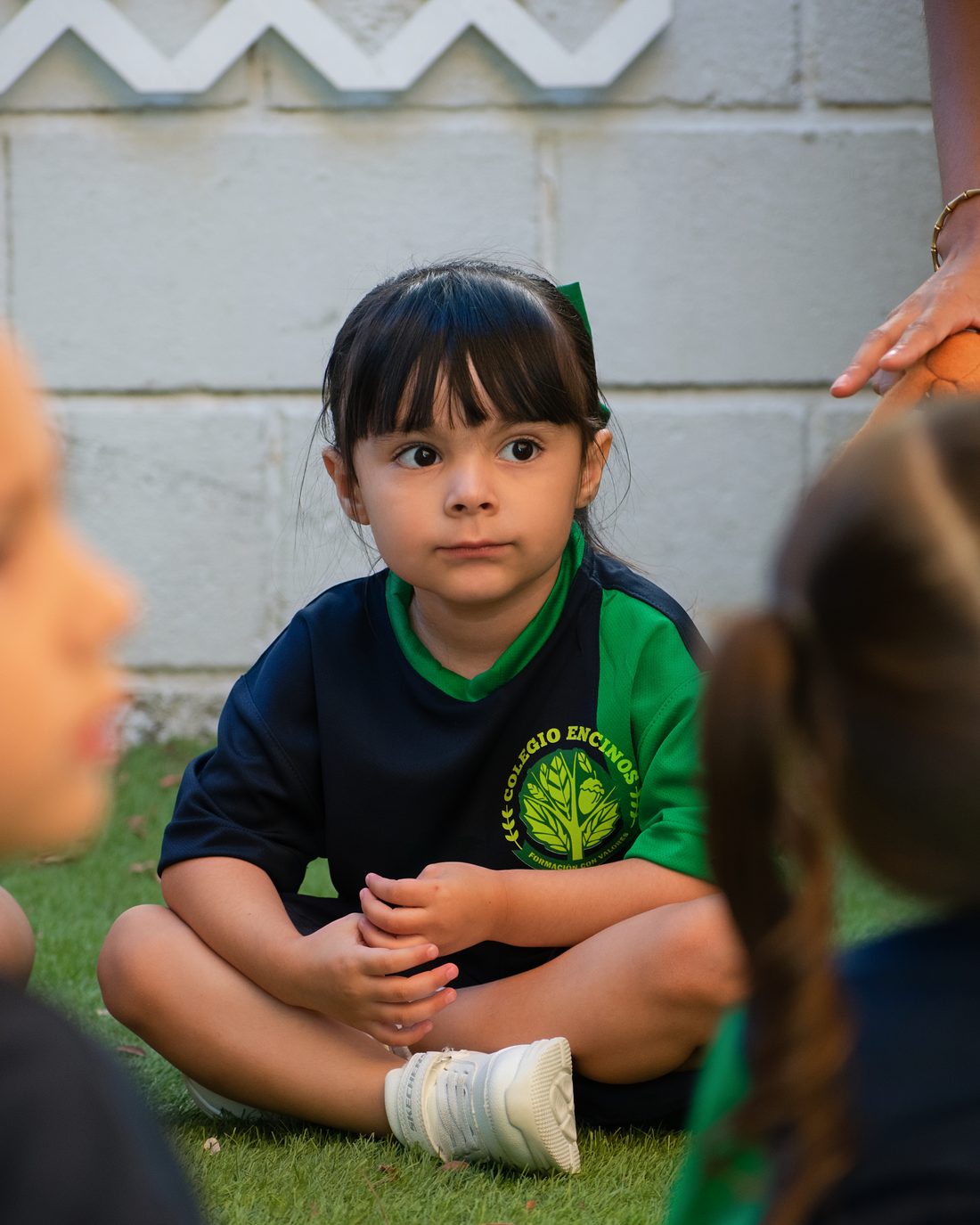 Niña de preescolar de Colegio Encinos sentada en el pasto, escuchando con atención a sus compañeros